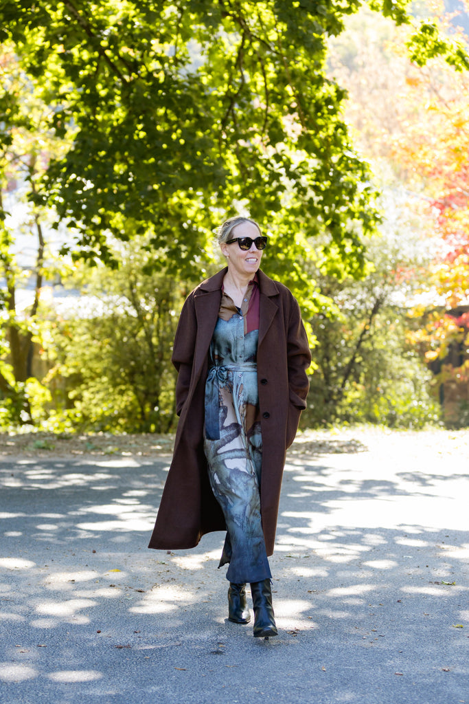 Woman standing in front of tree with filtered light and had on overcoat with dress and black boots.