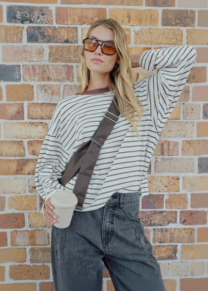 Woman wearing a striped sweater and sunglasses, holding a cup against a brick wall.
