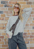 Woman wearing a striped sweater and sunglasses, holding a cup against a brick wall.