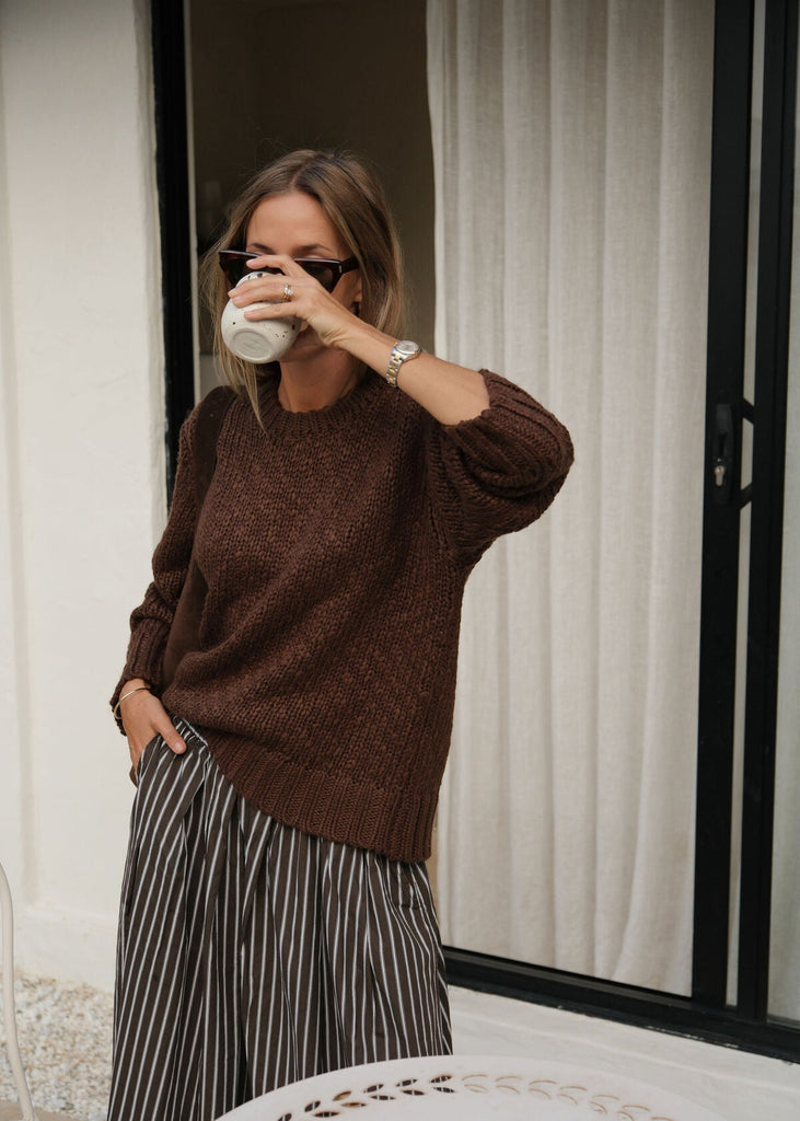 Woman in a brown sweater and striped skirt holding a white mug, standing in front of a mirror.