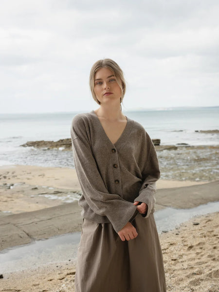 Woman standing on a beach wearing a long, light brown cardigan.