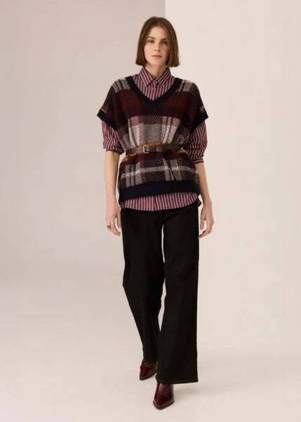 Woman wearing a patterned sweater vest over a striped shirt with black pants on a beige background